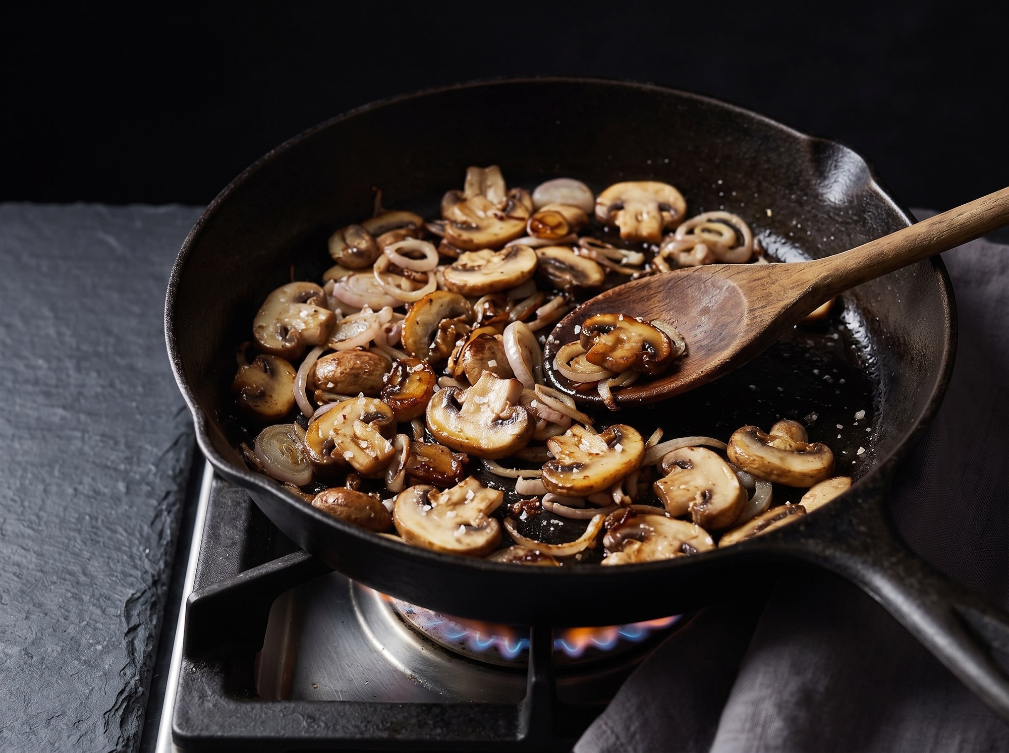A cast iron skillet on a gas stove holds sliced mushrooms and shallots browning with crisped edges, coarse salt scattered on top, a wooden spoon resting in the pan.